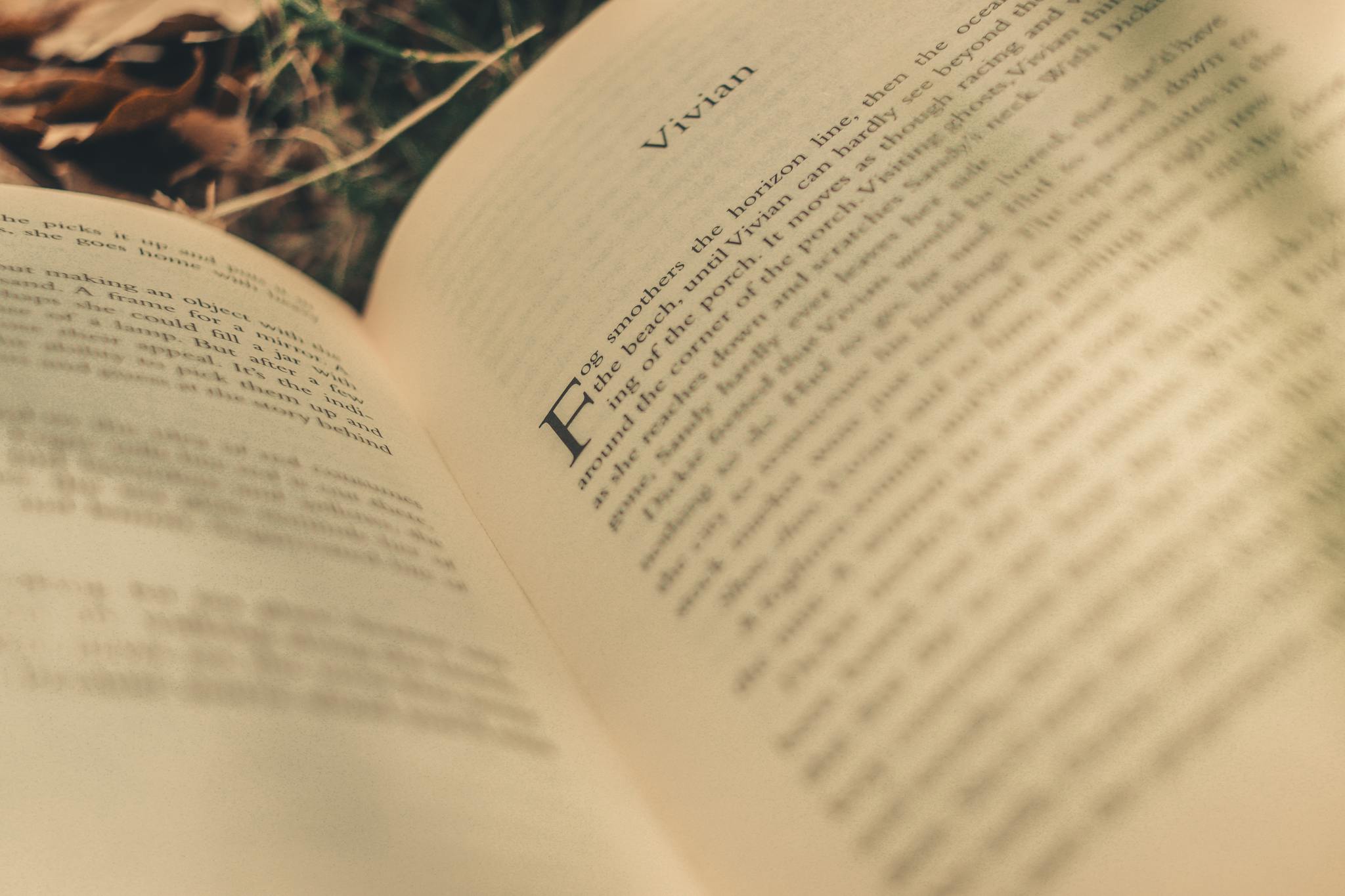 A close-up of an open book page with blurred text, surrounded by leaves, outdoors.