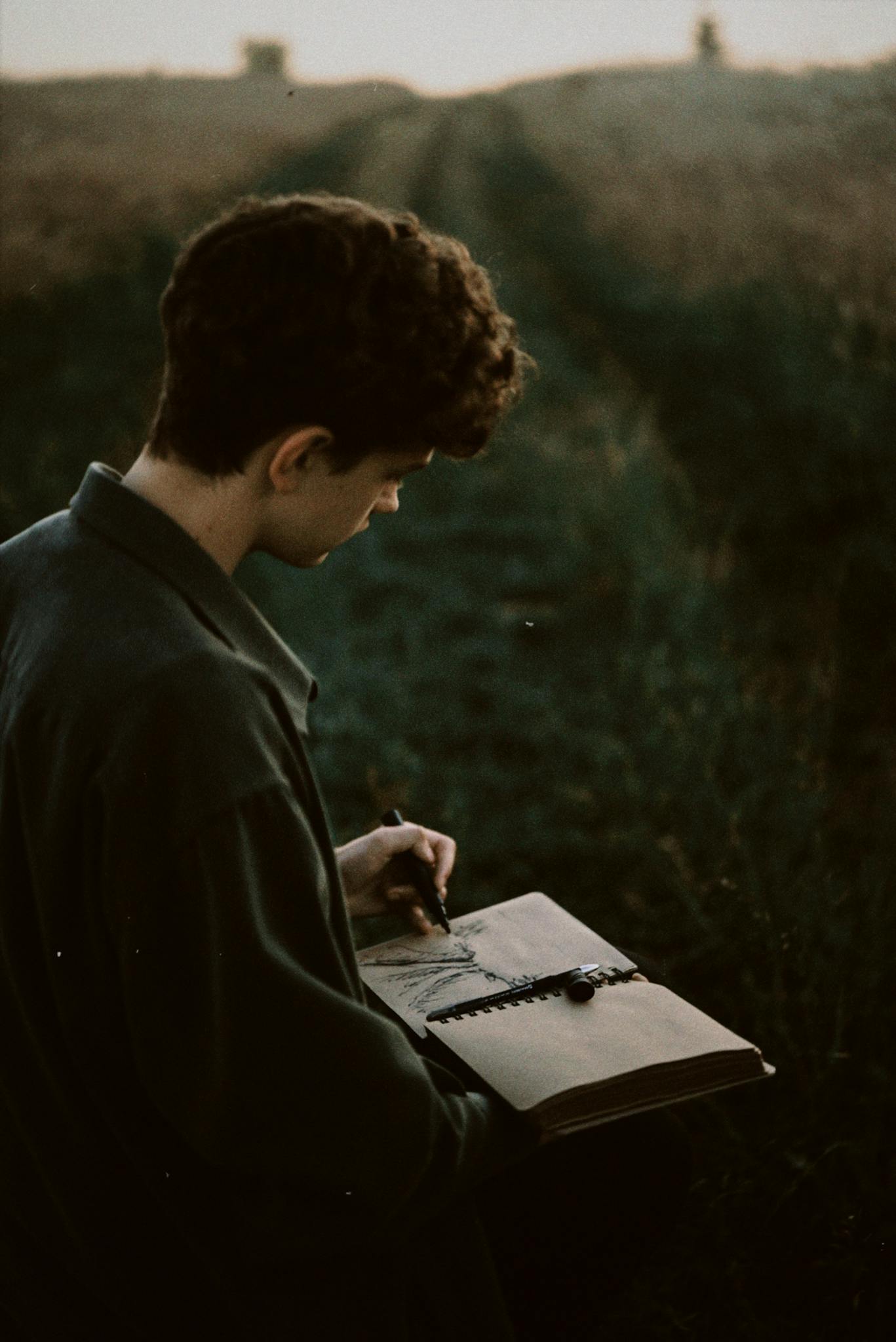 A young man writing in a notebook in a serene outdoor setting during twilight.