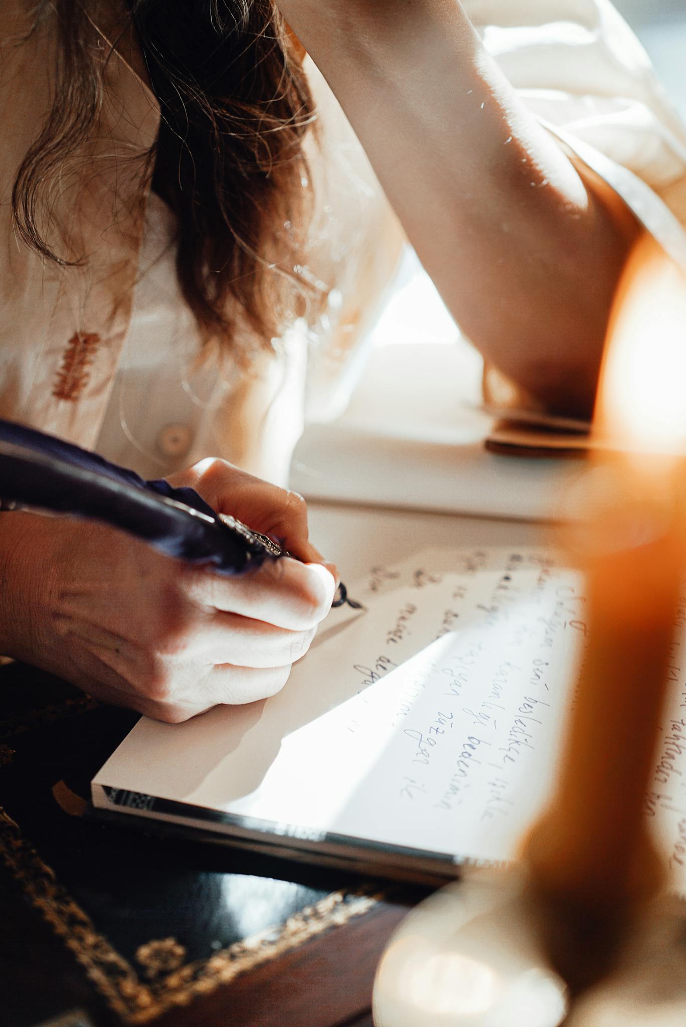Crop anonymous female writer with feather taking notes in notebook while leaning on hand in sun ray