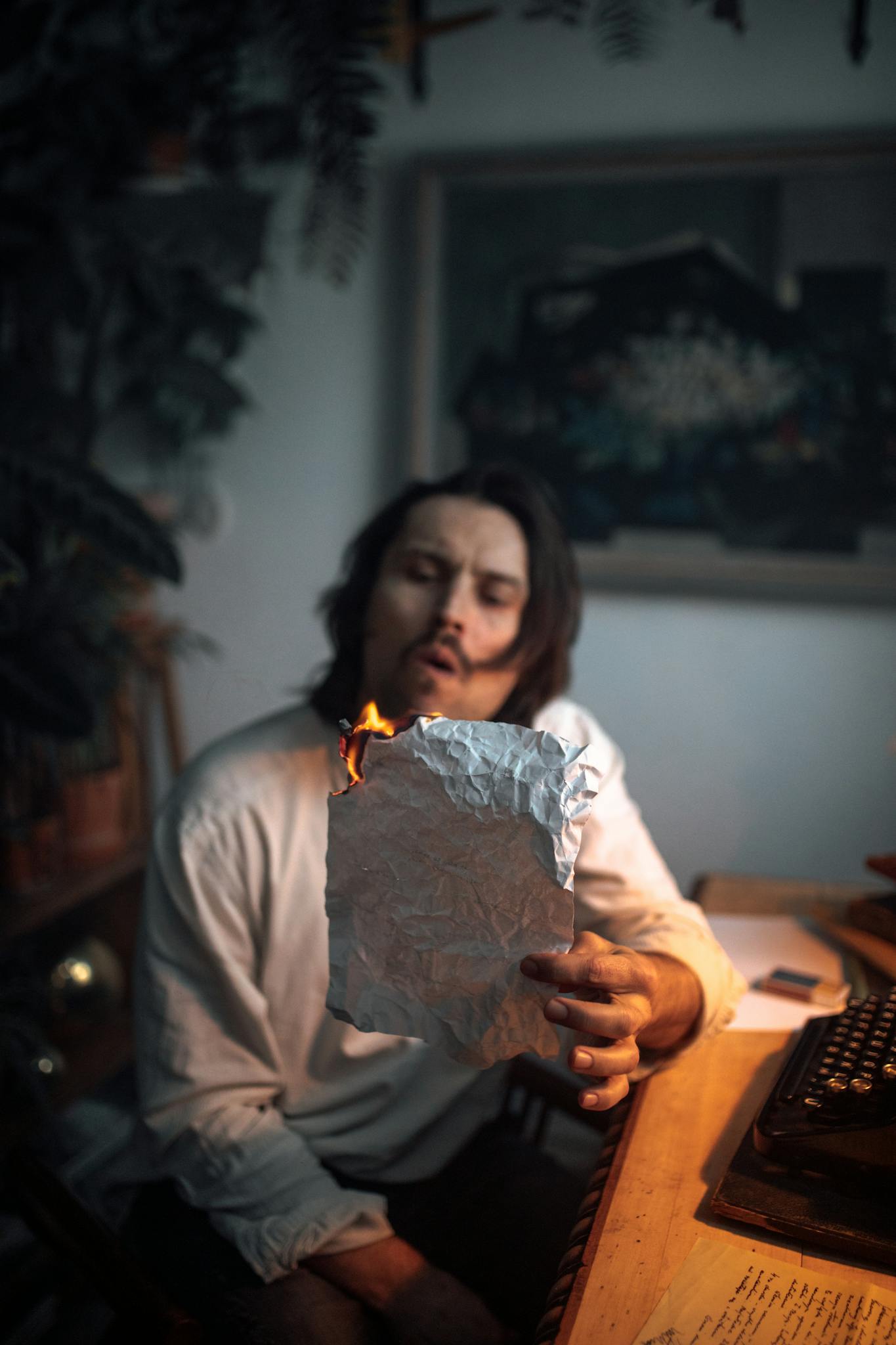 A man sits indoors blowing on a burning paper at a desk featuring a vintage typewriter, evoking creativity.