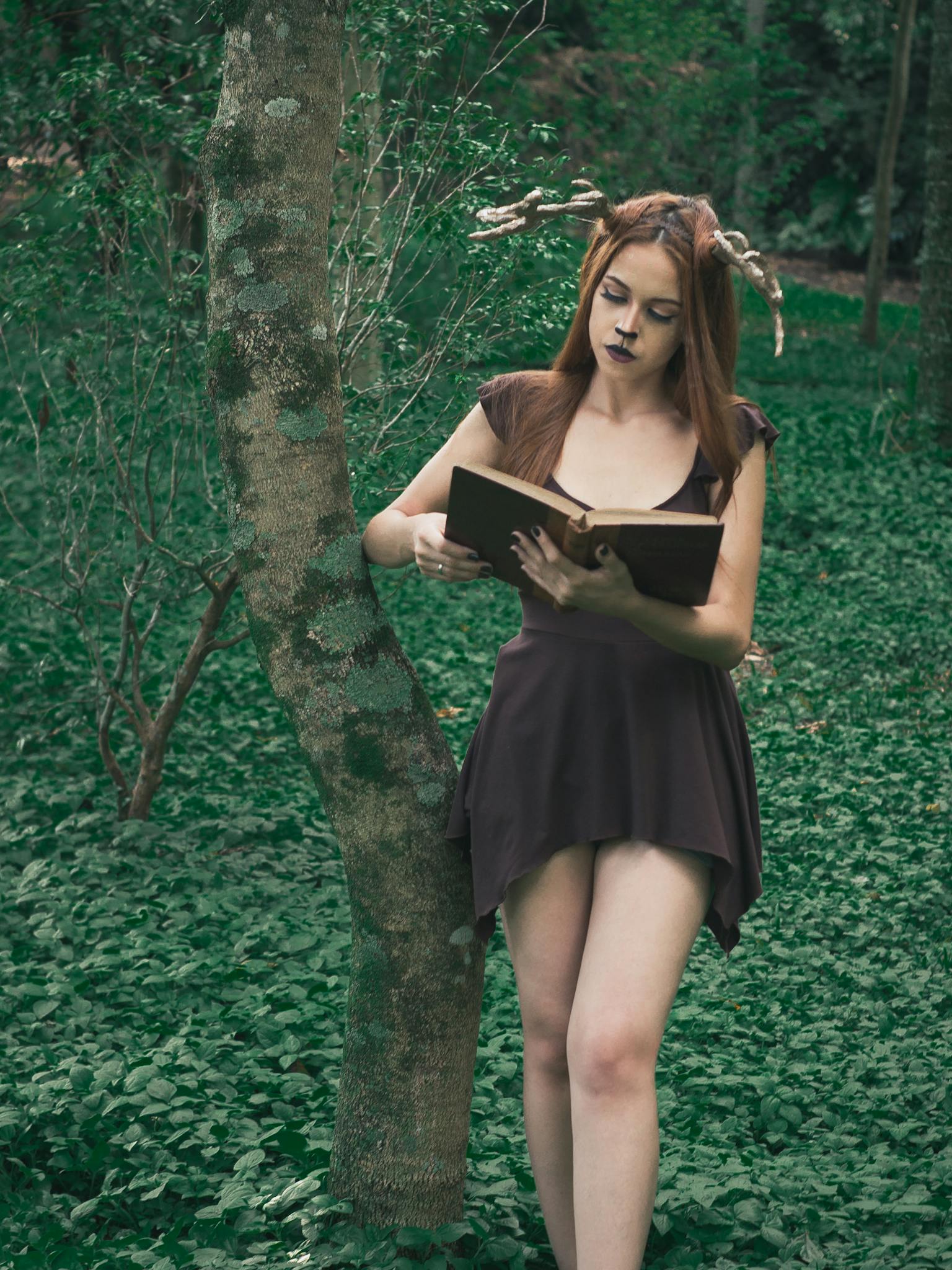 Young woman in a forest with deer antlers, reading a book, merging nature and fantasy.